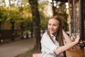 Woman blonde hair stands in front of a street coffee kiosk and is about to place an order. A woman fix her hair, look at side and waits for an order while walking in park.  © zvkate
