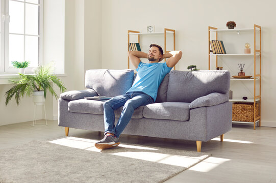 Young relaxed man sitting on comfortable couch in the living room at modern home and having a rest from a work with closed eyes. Calm peaceful guy enjoying quiet time alone in the room and dreaming.