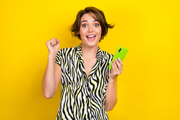 Portrait of overjoyed ecstatic woman wear striped blouse hold smartphone win betting clenching fist isolated on yellow color background