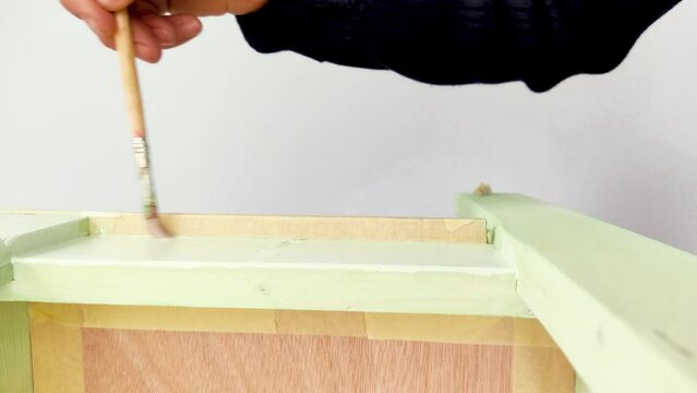 Close Up View of a Man's Hand Painting Pale Green Color a Wooden Chair with a Paintbrush Indoors.