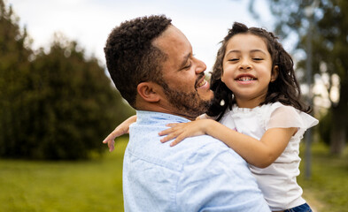 A dark-skinned man poses happily with his daughter in an outdoor park. The chubby African adult is holding the little girl looking at the camera. Concept of African-American parents.