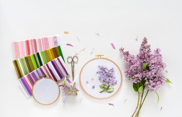 Summer needlework. Hand embroidery with satin ribbons of blooming lilacs in round wooden hoop, sets of colorful ribbons, accessories for embroidery on white background. Flat lay, copy space, close-up