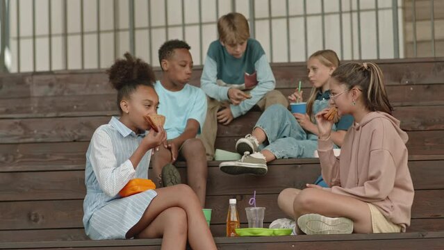 Multiethnic group of cheerful schoolkids sitting on outdoor stairs having lunch and chatting