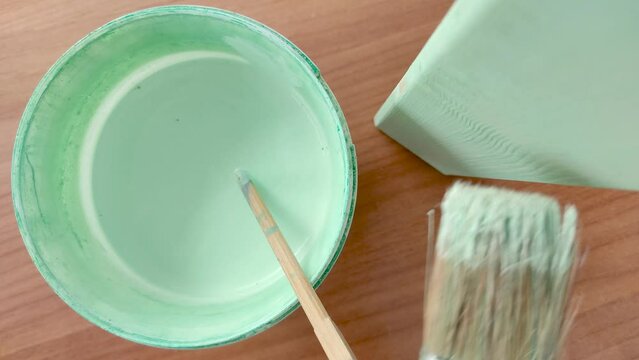 Hand of a Man Holding a Brush and Painting with Green Turquoise Color a Chair Leg on a Table. A Bucket with a Stirring Stick in it is on the Table.