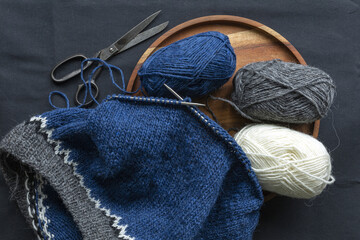 Fototapeta na wymiar Blue grey white wool knitted sweater on the metal needles with yarn balls and vintage scissors, stockinette stitch knitting in progress close-up photo