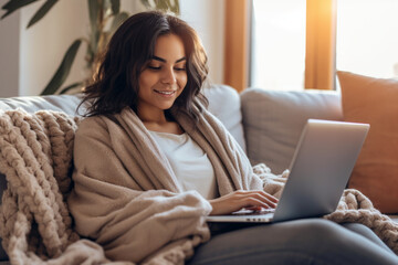 Ethnic woman sitting on a cozy couch, wrapped in a blanket, while working on her laptop, ethnic woman working, business, natural light, affinity, bright background Generative AI