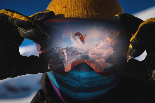 Close-up portrait of a skier in a mask holding on to ski goggles in the reflection of which the skier does a backflip trick against the backdrop of snowy epic high mountains