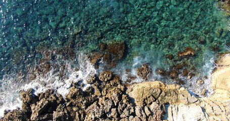 Aerial top view over the island's rocky shore and clear sea with varying shades of blue and light foamy waves in warm summer weather.