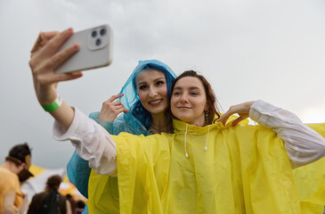 Happy queer couple taking a selfie on a summer festival in the rain. Cheerful LGBT females wearing...