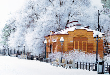 Naklejka premium Snow-covered red brick public toilet and tree branches in fantastic hoarfrost. Winter landscape with benches and lanterns in a city park after a snowfall. No people.