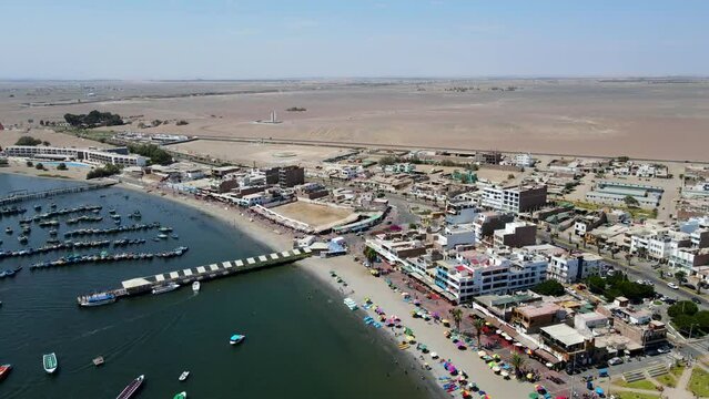 Aerial shot drone orbits to the right over boats in the harbor and sumbrellas on the beach in the desert town of Paracas, Peru