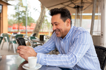 Happy mature latin man using cell phone sitting at street cafe
