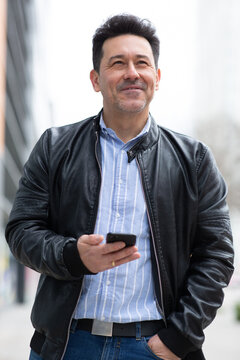 Happy older latin man in black leather jacket with cellphone standing outside