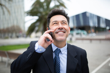 Smiling mature businessman standing outside in city and talking on mobile phone