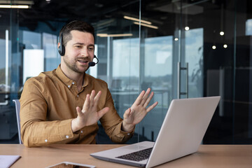 Fototapeta na wymiar Online consultation, hotline support service. A young man sits in the office in a headset and talks on a video call with clients. Gestures with hands, calms, advises