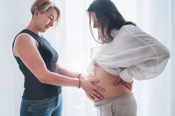 Young woman tender touching partner's female pregnant belly. Same-sex marriage couple next to...