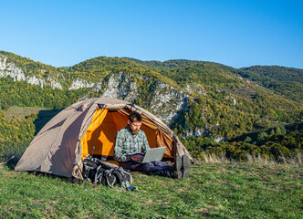 The concept of remote work or freelancer lifestyle. Young man using computer notebook to check news in front of a tent while camping on a holiday