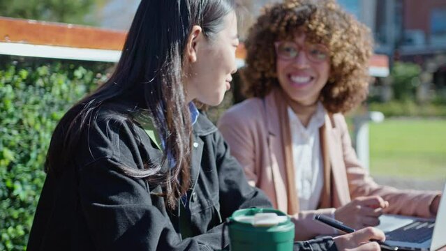 Young African-American and Asian female colleagues working with laptop, discussing business plans, sitting together on street bench outdoors in the city. Medium rack focus shot