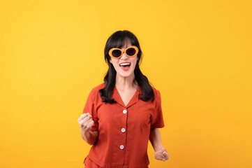 Portrait young beautiful asian woman happy smile dressed in orange clothes showing fist up hand gesture isolated on yellow studio background. Successful hooray celebrate young person concept.