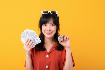 Portrait young beautiful asian woman happy smile dressed in orange clothes holding cash money and crypto bitcoin digital currency celebrate her success isolated on yellow background. Be rich concept.