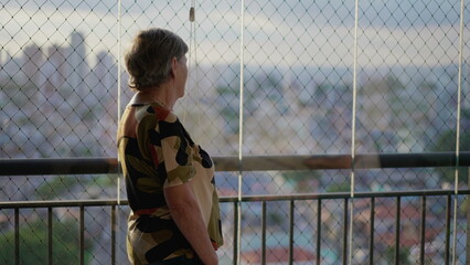 Older retired woman standing at apartment balcony looking at city view. Senior female person in 70s overlooking cityscape