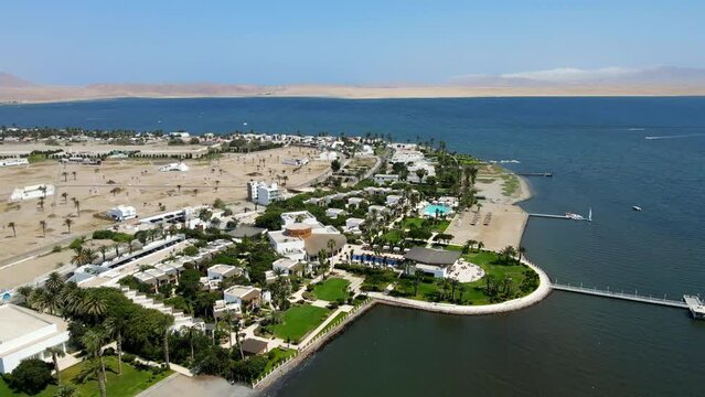Aerial shot drone orbits to right around beachside resort clashing with the desert landscape and the Paracas National Reserve in the background