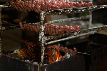 Raw sausage italian pork sausage salsiccia is grilling on barbeque grill with fire by summer night outdoor. Freshly made raw breed butchers sausages is grilling. Street food