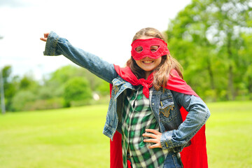 Girl Plays Superhero. Happy girl are playing superhero. Kid run across green field in red cloak at...