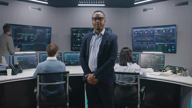 African American IT scientist stands in monitoring room and looks at camera. Multi cultural big data engineers work at computers. Multiple screens with displayed real-time analysis charts. Dolly shot.