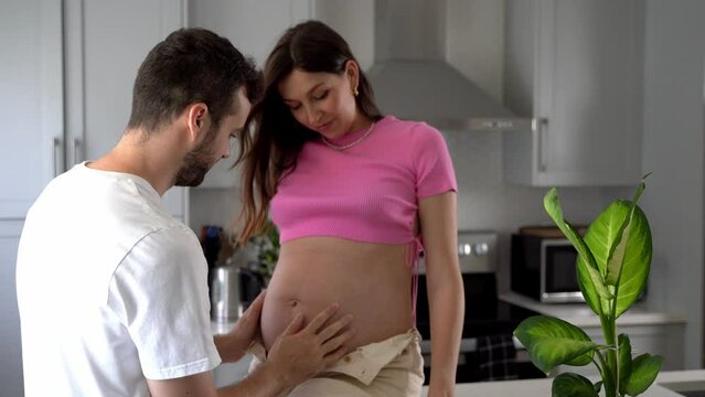 Young Caucasian couple future parents together smiling in kitchen and husband caresses the belly of his pregnant wife. Family expecting baby hugging each other at home. Husband and wife