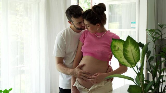 Young Caucasian couple future parents together smiling in kitchen and husband caresses the belly of his pregnant wife. Family expecting baby hugging each other at home. Husband and wife
