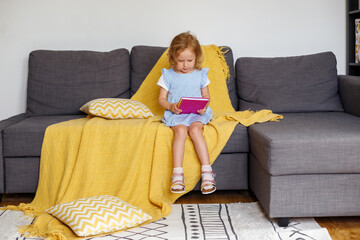 Little girl sitting on sofa and touching a notepad. Child suffering from stomach ache at home.