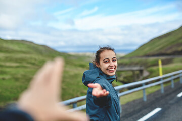 Fototapeta premium Young laughing girl outstretching hand asking to follow her on background of mountain road, Iceland, West Fjords.