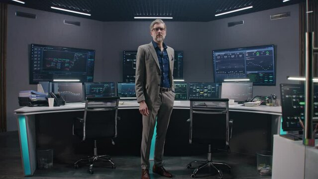 Mature big data scientist in business suit puts on glasses, stands in monitoring room and looks at camera. Multiple big screens on the wall with displayed live analysis feed at background. Dolly shot.