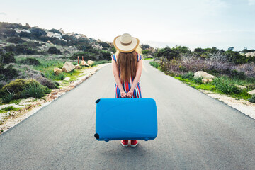 Beautiful woman walking on the road in hat holding suitcase. Back view, travel concept