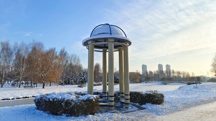 In a snow-covered city park, a rotunda of concrete columns with a dome-shaped roof stands next to a pedestrian path and an ice-covered canal. Behind the park's trees stand buildings. Sunny and frosty