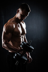 Fototapeta na wymiar Athletic shirtless young male fitness model makes exercises with dumbbells on dark background