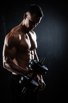 Athletic shirtless young male fitness model makes exercises with dumbbells on dark background