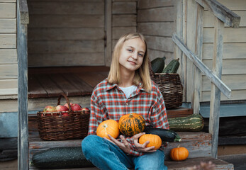 Girl is sitting on steps of an old village house with pumpkin in hands near sunflowers and baskets with zucchini and apples. Harvest. Autumn. Fall. Halloween. Thanksgiving day