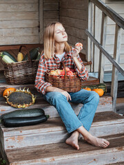 Girl is sitting on steps of an old village house with her face exposed to warm sunbeams, holding large basket with apples on her knees and eating apple. Ripe pumpkins and sunflower lie nearby. Harvest