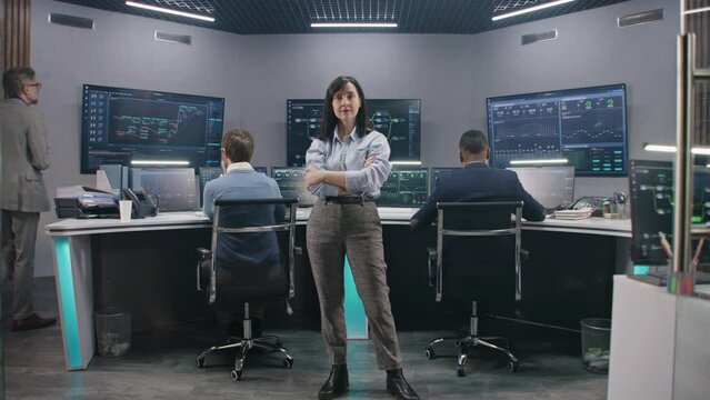 Female big data scientist stands in monitoring room and looks at camera. Software engineers work at computers at background. Multiple big screens with displayed real-time analysis charts. Dolly shot.