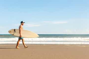 Fototapeta na wymiar Handsome fit man walk with white blank surfboard wait for wave to surf spot at sea ocean beach. View from side. Concept of sport, fitness, freedom, happiness, new modern life, hipster, generation Y.
