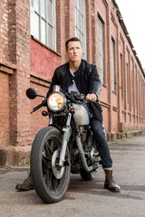 Handsome rider biker guy in leather jacket sit on classic style cafe racer motorcycle and look...