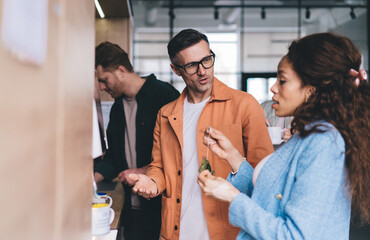 Caucasian man in optical spectacles for vision protection communicate with blurred African American woman during working process in office, diverse male and female colleagues talking at kitchenette