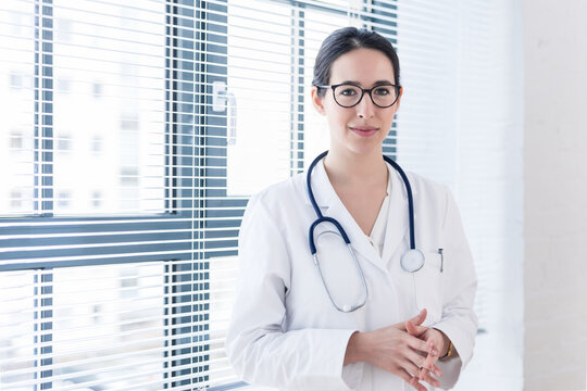 Portrait of a young nurse or physician wearing eyeglasses and white medical gown while looking at camera with confidence and serenity indoors