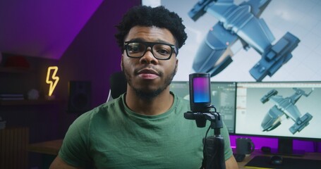 African American 3D designer speaks on camera using microphone, records video tutorial about 3D modeling and animation. 3D aircraft design and software interface on computer and big digital screen.