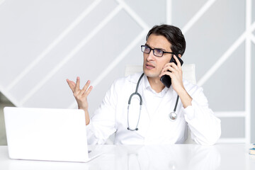 Close up portrait of tired male doctor sitting at the desktop and talking to phone in the office of modern clinic	