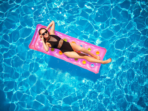 Fit Pretty Girl In Bikini Chilling On Inflatable Pink Mattress In Swimming Pool. Slim Hot Woman In Swimwear Tanning. Female Relaxing On Float In Blue Water At Luxury Resort. Aerial, View From Above.