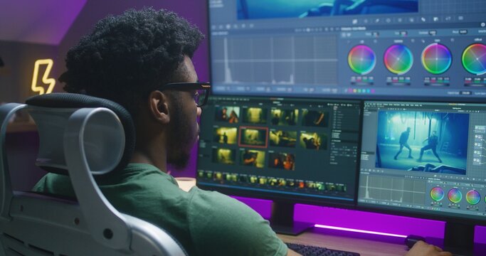 African American video editor makes color grading, works at home office. Action movie footage and program interface on multi-monitor computer and big digital screen. Concept of film post production.