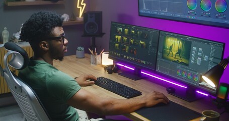 African American editor makes color correction, works at home studio. Action movie footage and program interface on multi-monitor computer and big digital screen. Concept of film post production.